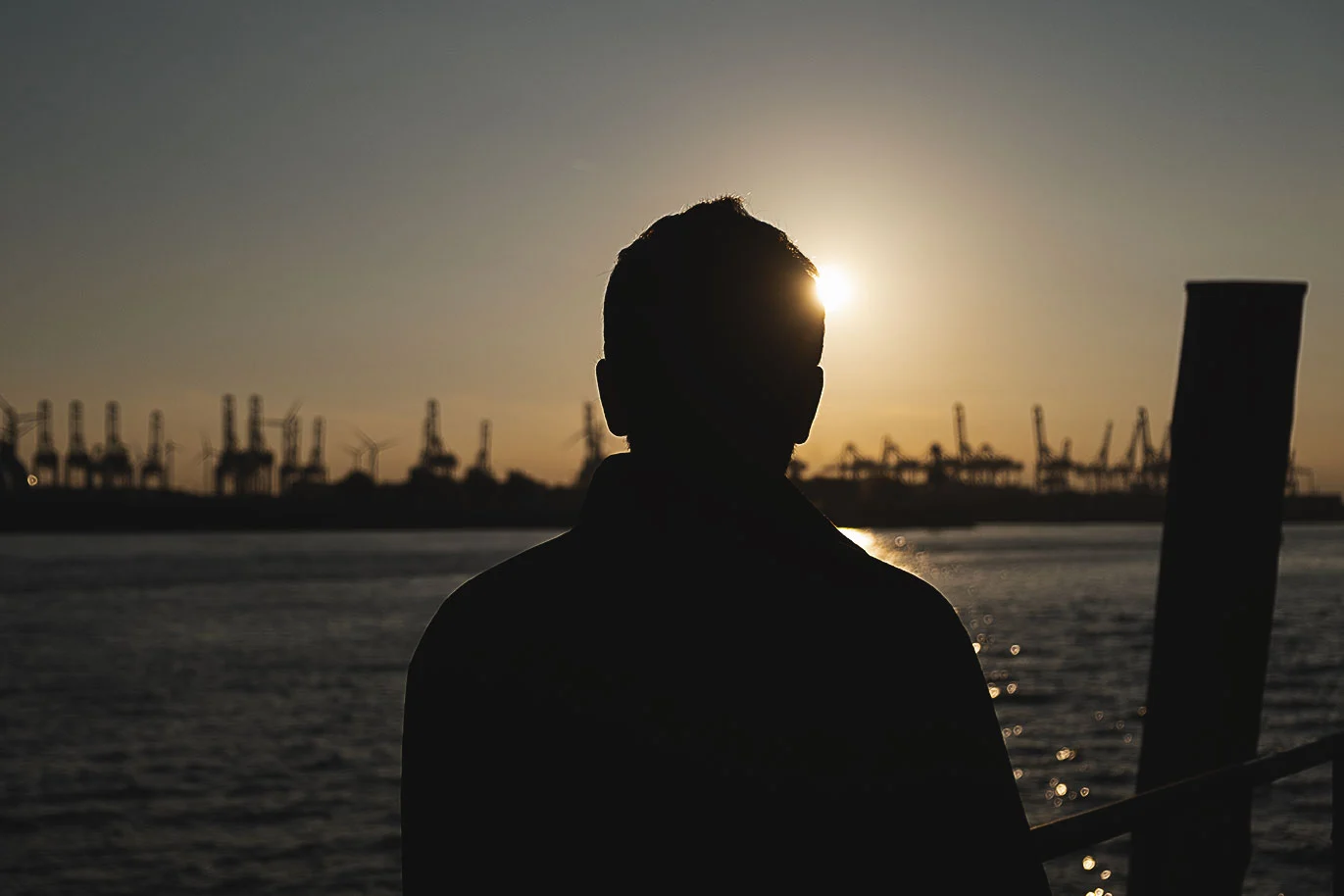 Silhouette eines Mannes, der im Sonnenuntergang am Hamburger Hafen steht, den Blick auf das Wasser gerichtet.