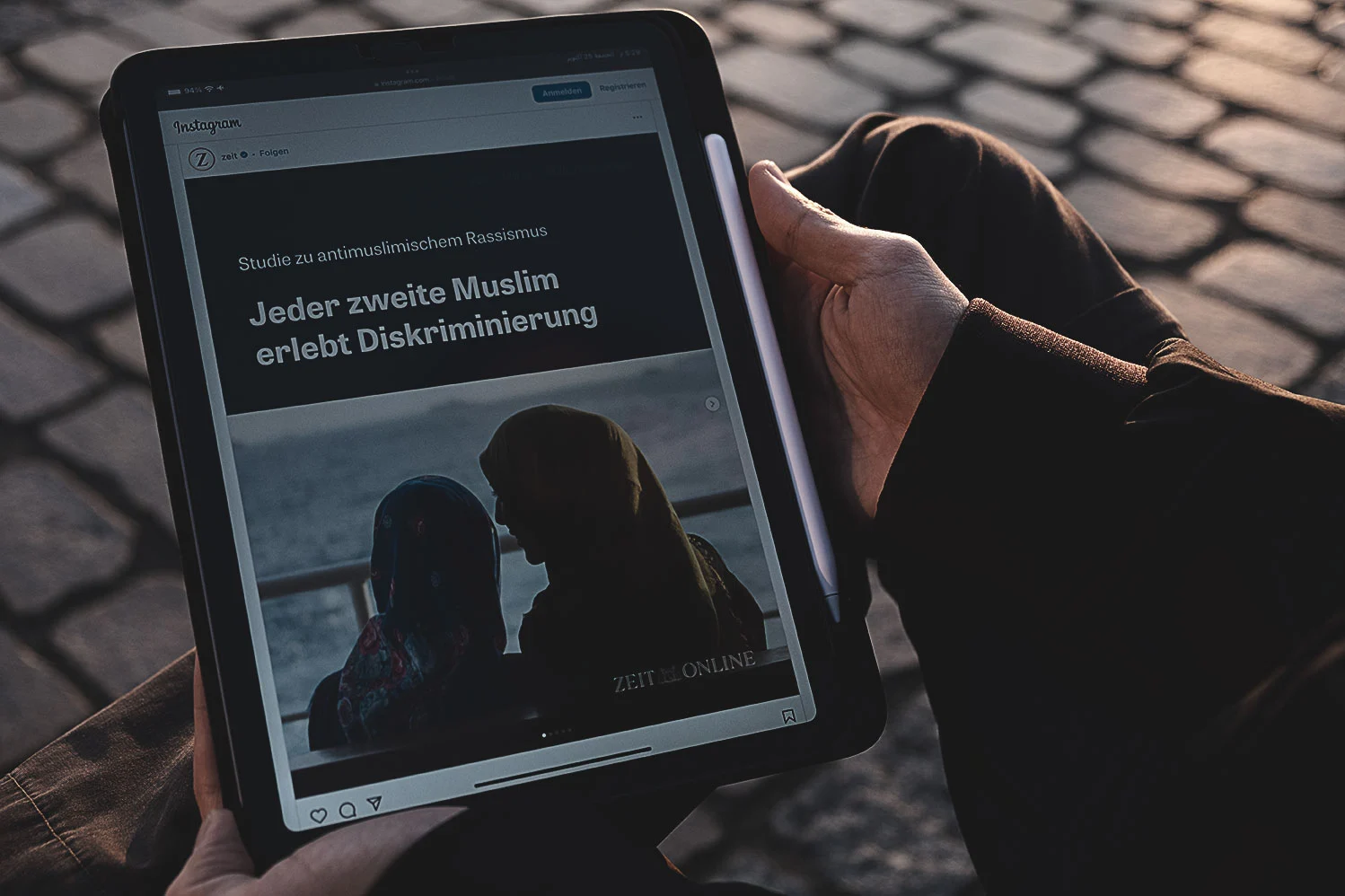Eine Person hält ein iPad, auf dem ein Instagram-Post von „Zeit Online“ zu sehen ist. Der Text auf dem Bildschirm lautet „Studie zu antimuslimischem Rassismus – Jeder zweite Muslim erlebt Diskriminierung“. Im Hintergrund ist das Bild von zwei Frauen mit Kopftuch zu erkennen.