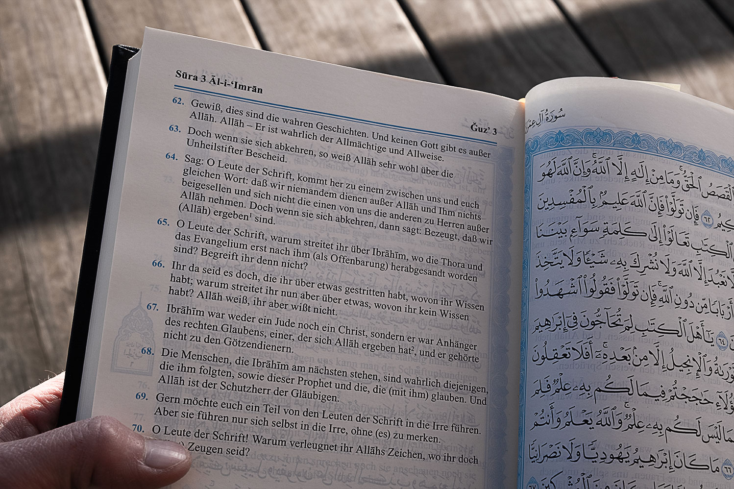 Eine Hand hält den Koran aufgeschlagen, auf der Seite von Sura 3, Al-‘Imran. Der arabische Text ist rechts abgebildet, während die Übersetzung der Bedeutung in die deutsche Sprache auf der linken Seite ist.