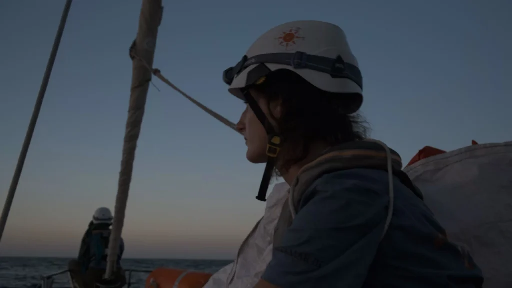 Eine Frau mit Helm, Stirnlampe und Rettungsweste schaut in die Ferne auf einem Boot im Abendlicht.