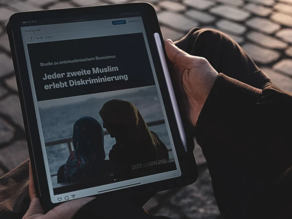 Eine Person hält ein iPad, auf dem ein Instagram-Post von „Zeit Online“ zu sehen ist. Der Text auf dem Bildschirm lautet „Studie zu antimuslimischem Rassismus – Jeder zweite Muslim erlebt Diskriminierung“. Im Hintergrund ist das Bild von zwei Frauen mit Kopftuch zu erkennen.