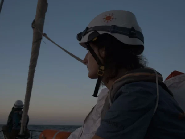 Eine Frau mit Helm, Stirnlampe und Rettungsweste schaut in die Ferne auf einem Boot im Abendlicht.